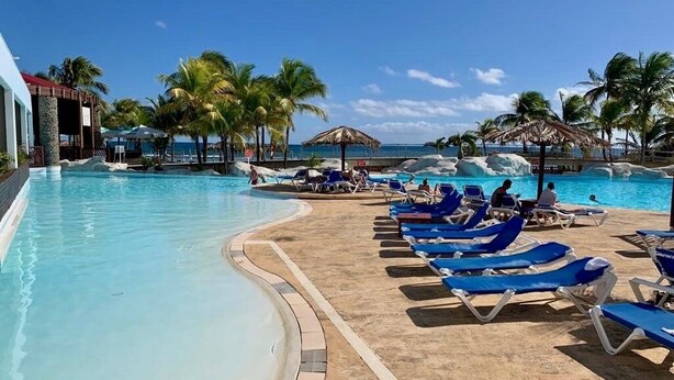 A resort pool area with clear blue water, lounge chairs, palm trees, and a view of the ocean.