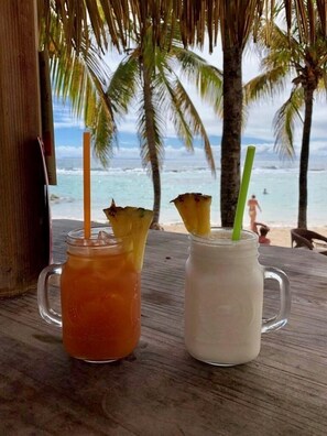 Duas bebidas com canudos sobre uma mesa de madeira, com vista para a praia e palmeiras ao fundo.