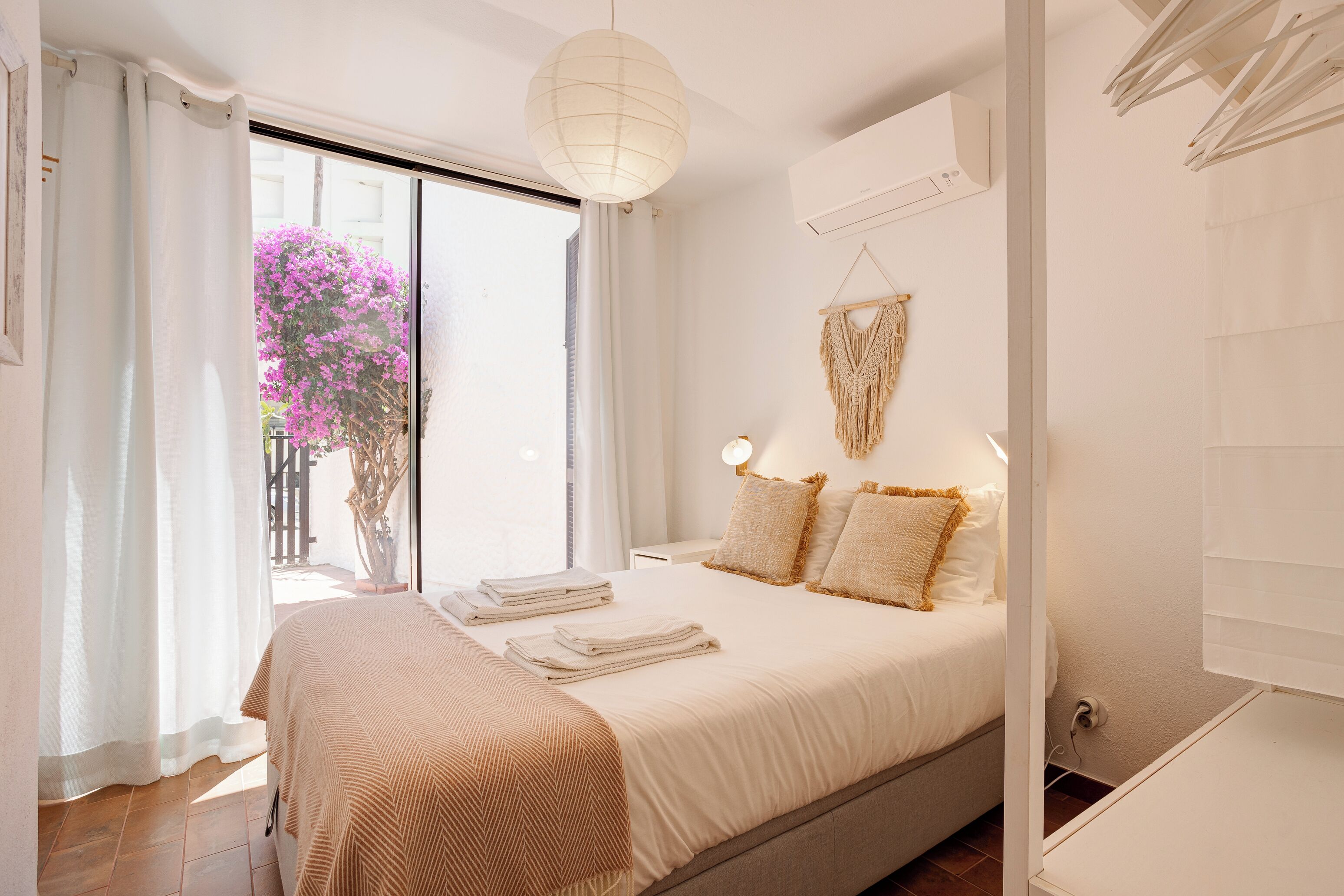Slaapkamer met een bed, gordijnen, een macramé wanddecoratie en een raam met uitzicht op roze bloemen.