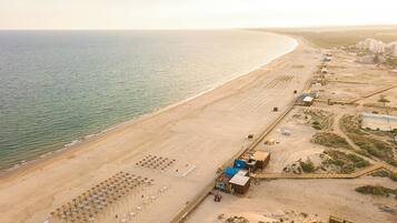 An aerial view of a beach with rows of umbrellas and sun loungers, a small building, and a distant shoreline.