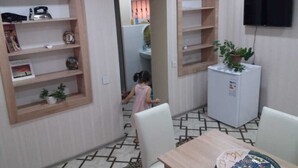 Un comedor con una mesa, sillas y un refrigerador. Hay estantes con diversos objetos y un televisor montado en la pared.