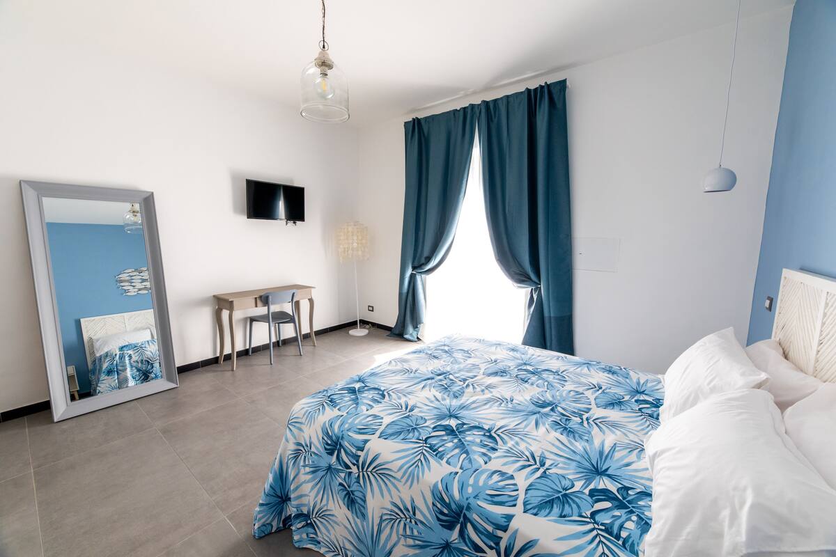 Une chambre à coucher comprenant un lit, un bureau, une chaise, un téléviseur et une grande fenêtre avec des rideaux.