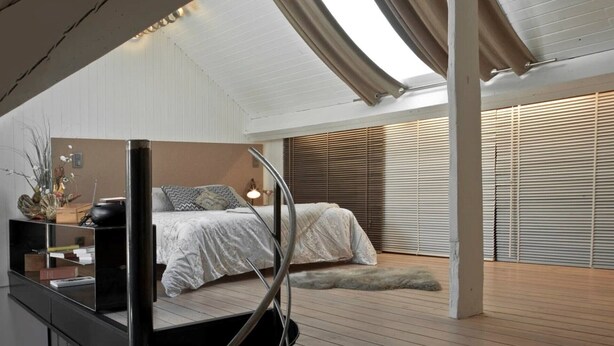 A modern bedroom with a bed, bedside table, lamp, and a view of the outside through large windows.