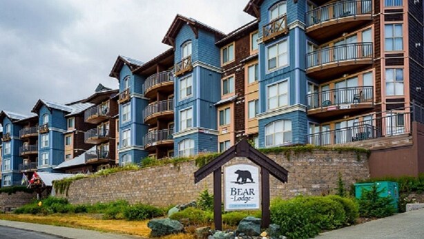 Une rangée d’immeubles à étages avec des balcons, une enseigne indiquant « Bear Lodge », et un mur de pierre orné de verdure.