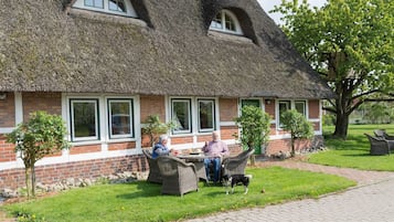 Ein Gebäude mit einem Reetdach und einer Ziegelverkleidung, mit Außenplätzen und einem kleinen Baum im Vordergrund.