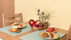 Une table en bois surmontée d’une nappe bleu clair, deux assiettes avec des croissants, un panier de pommes et un vase rempli de fleurs.
