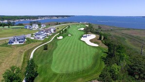 An aerial view of a golf course with a clubhouse, sand bunkers, and a body of water in the background.