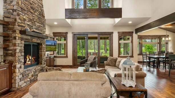 A spacious living room with a stone fireplace, large windows, and a high ceiling.
