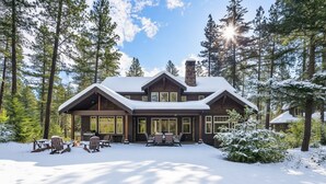 A large, two-story log cabin with a snow-covered roof, surrounded by tall pine trees.