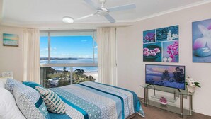 A bedroom with a large window offering a view of the sea, a bed with patterned bedding, a ceiling fan, and a glass TV stand with a painting. - Ocean Plaza 1467 - Coolangatta Beachfront - Min. 3 Night Stays! (Coolangatta)