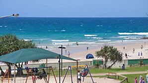 A beach with a swing set, a shaded area, and a lifeguard stand. - Ocean Plaza 1467 - Coolangatta Beachfront - Min. 3 Night Stays! (Coolangatta)