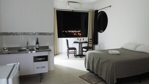 Ein kompaktes Hotelzimmer mit Küchenzeile, einem Bett mit kariertem Überwurf, einer Essecke mit Tisch und Stühlen sowie Blick auf die beleuchtete Stadt.