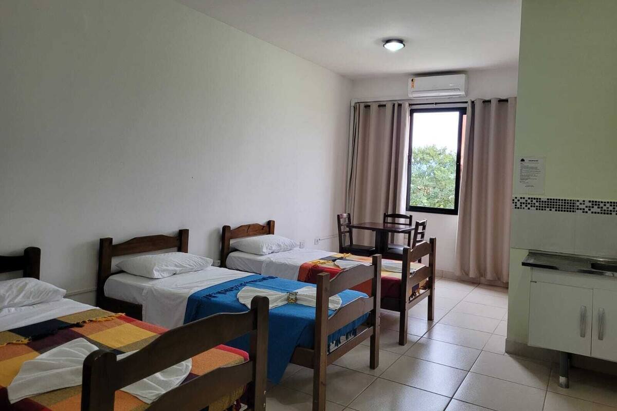 Une chambre d’hôtel avec deux lits simples, une petite table avec des chaises, une fenêtre avec des rideaux et un coin cuisine.