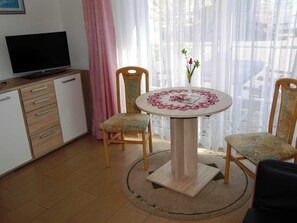 Een kleine, goed verlichte kamer met een televisie op een kastje, twee houten stoelen rond een ronde tafel met een decoratieve tafelkleed, en een raam met dunne gordijnen.