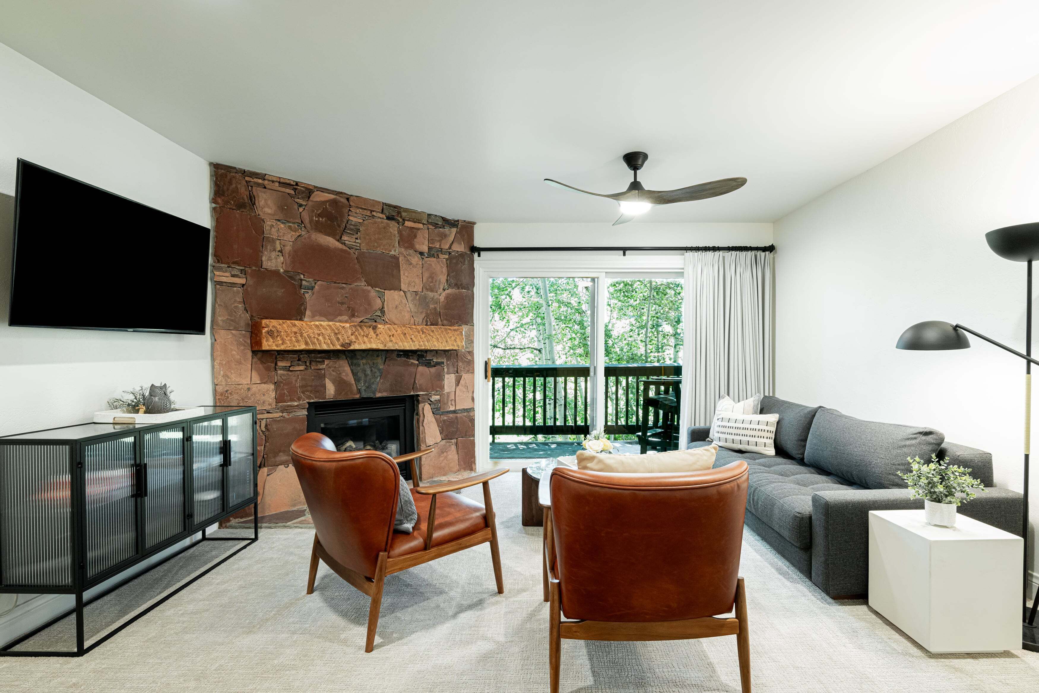 Un salón con chimenea de piedra, televisor de pantalla plana, sillones de cuero, sofá gris y balcón con vistas a árboles.