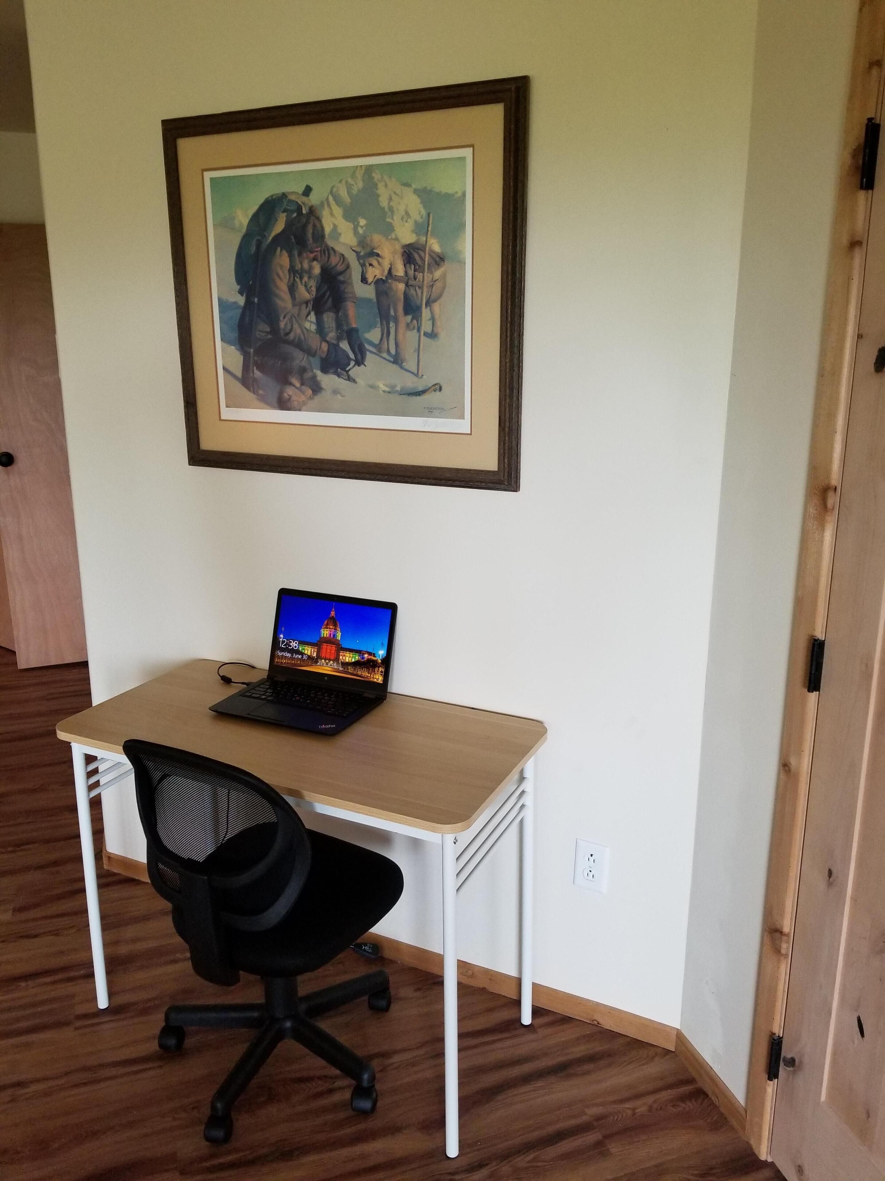 Une pièce avec un bureau en bois, un fauteuil de bureau noir et un tableau encadré accroché au mur.