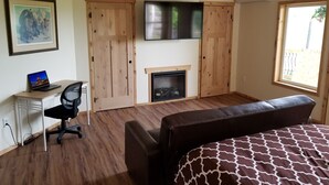 A bedroom with a bed, a desk with a laptop, a fireplace, and a TV mounted on the wall. - Fort Wrangell, Orca Room (Wrangell)