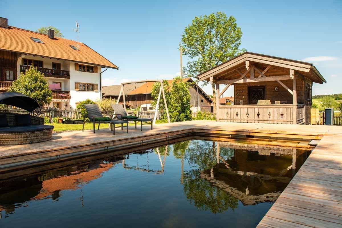 A swimming pool with a wooden deck, a covered seating area, and a building with a sloped roof.