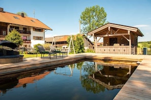 Een zwembad met een houten terras, een overdekte zithoek en een gebouw met een schuin dak.