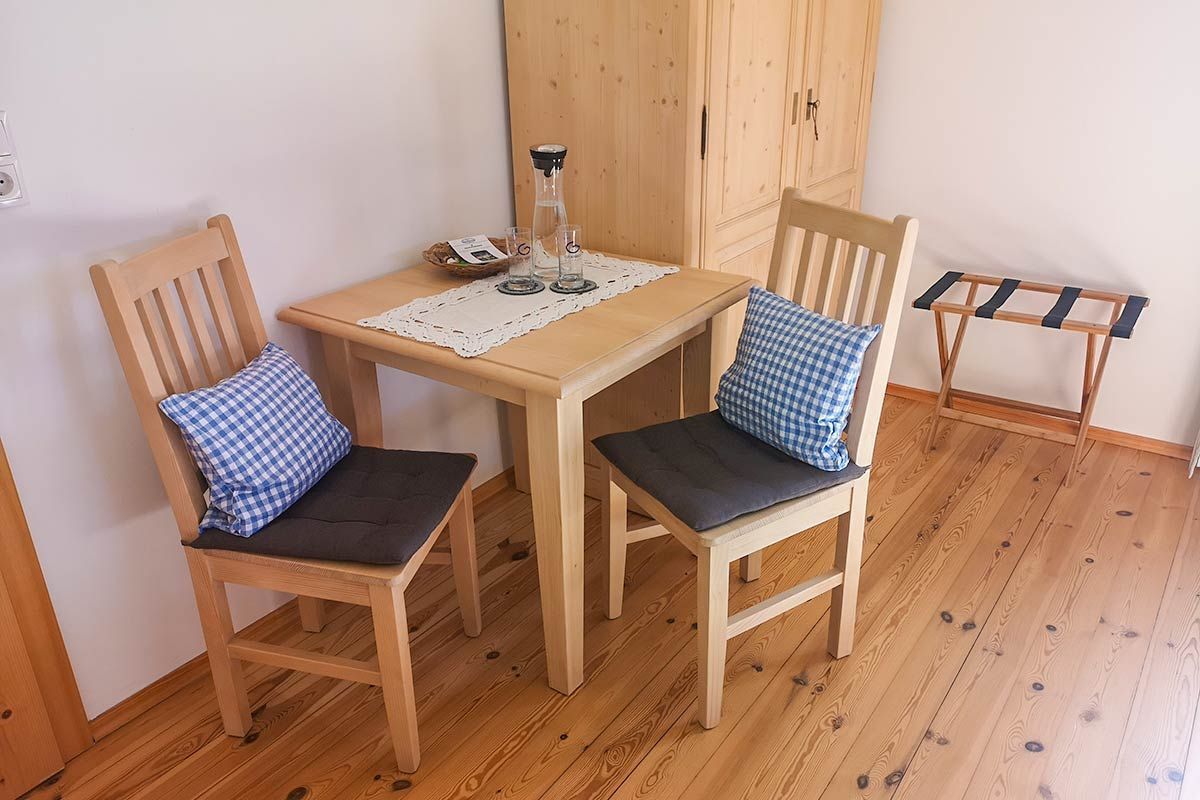 Une petite pièce avec deux chaises en bois, une table recouverte d’une nappe en dentelle, une bouilloire et un étendoir.