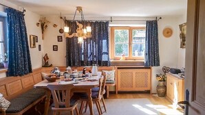 Un comedor acogedor con una mesa de madera, sillas y un banco. Hay una ventana con cortinas, una araña y un reloj en la pared.