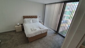 A bedroom with a large bed, a nightstand, a lamp, and a window with blinds.