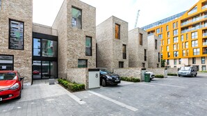 A modern building with a brick facade, a glass entrance, and a parking area with a red car and a blue van.