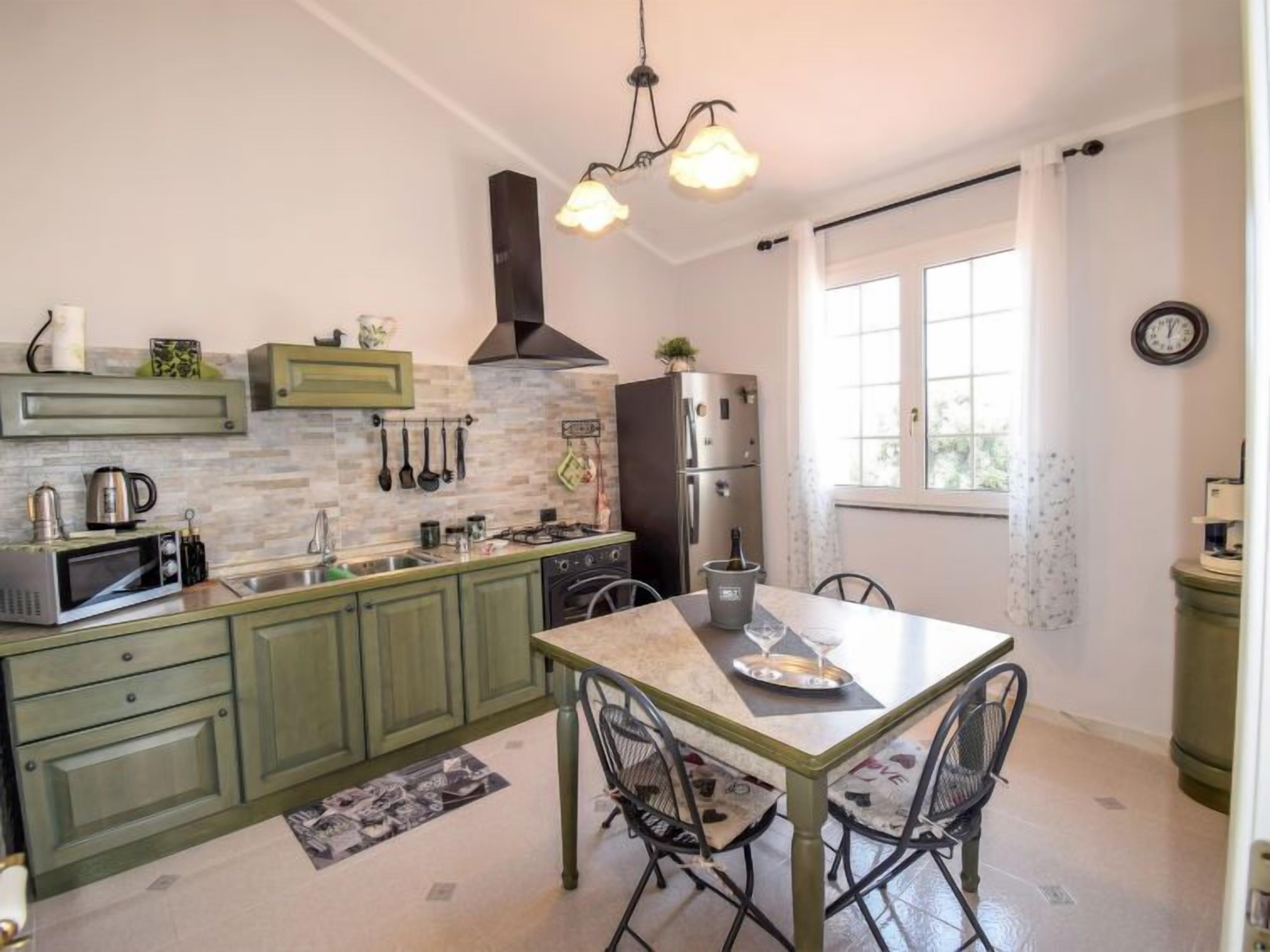 A kitchen with green cabinets, a stainless steel refrigerator, a microwave, and a gas stove. There is a dining table with chairs and a chandelier above.