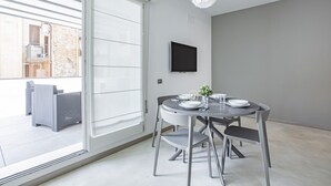 Un área de comedor moderna con una mesa redonda, sillas grises, un televisor de pantalla plana montado en la pared y vistas al balcón exterior.