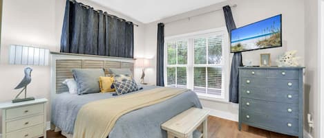 A bedroom with a bed, bedside table, bench, dresser, TV, window, curtains, and ceiling fan.