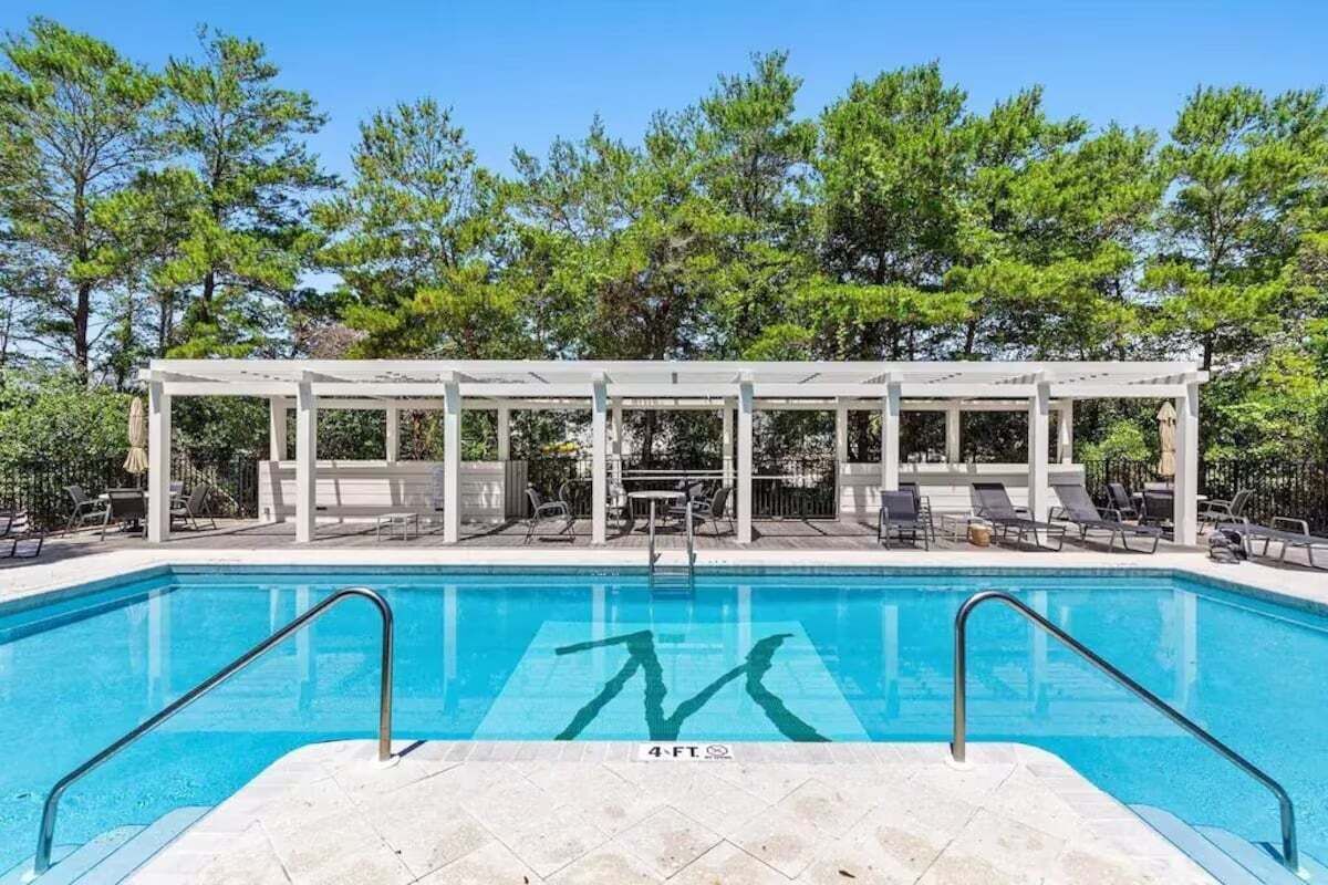 A rectangular swimming pool with a white pergola structure in the background, surrounded by trees.