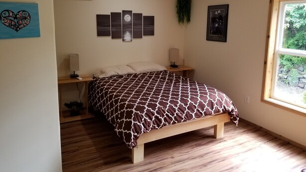 Een slaapkamer met een bed, nachtkastjes, een wandairconditioner, een raam met uitzicht op groen, en een ingelijste foto aan de muur.