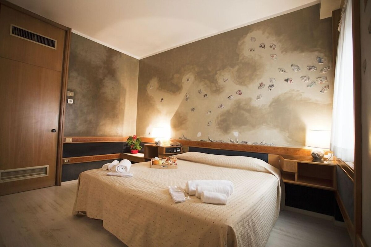 A hotel room with a large bed, two bedside tables, a wooden wardrobe, and a wall with a bird mural.