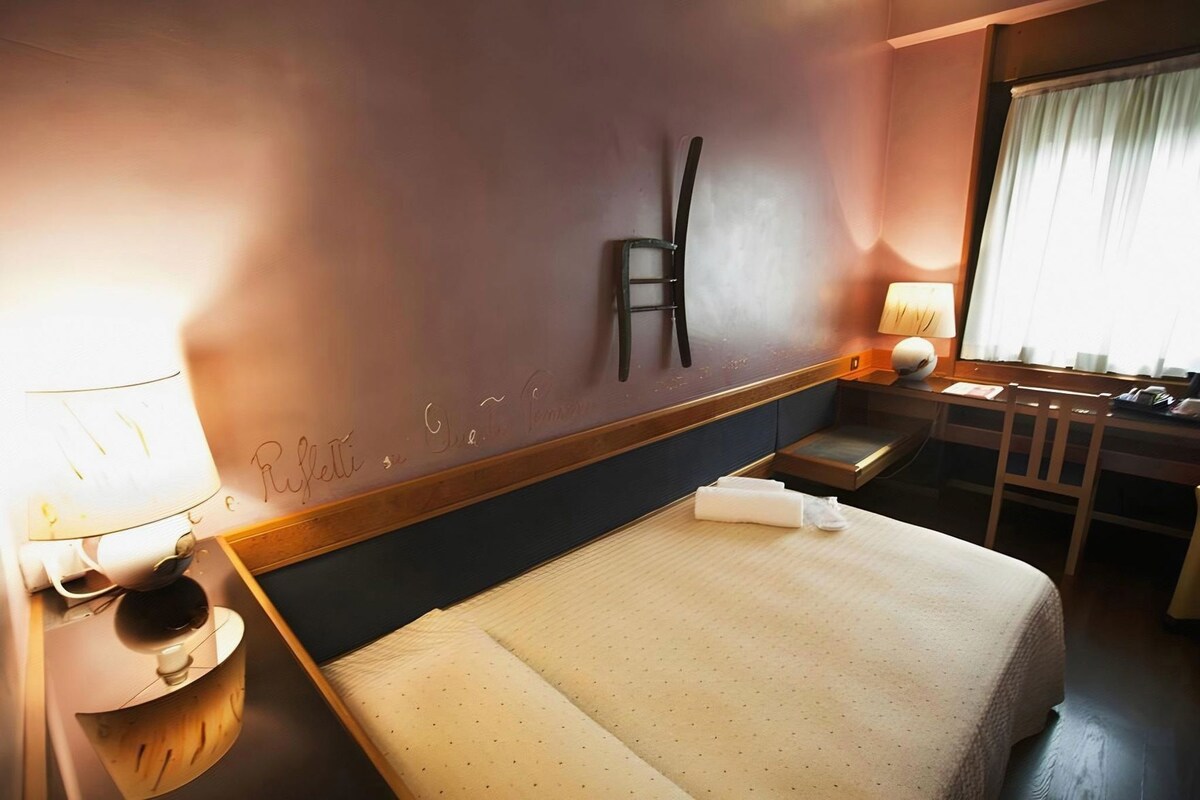 A hotel room with a bed, bedside table, lamp, chair, and a desk with a chair.