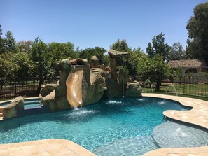 A swimming pool with a rocky waterfall feature, surrounded by a fence and trees. - Oasis Getaway in Gilbert AZ Perfect for the family (Gilbert)