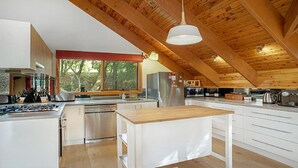 Une cuisine moderne avec des poutres en bois, un grand îlot central, des appareils électroménagers en acier inoxydable et une vue sur les arbres à l’extérieur.