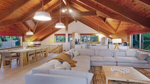 Un loft spacieux avec de hauts plafonds, des poutres apparentes, un coin repas, un salon avec un canapé et une table basse, et de grandes fenêtres offrant une vue sur l’extérieur.