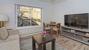 A living room with a sofa, a coffee table with a vase, a TV on a stand, a dining table with chairs, and a large window with a view of a staircase. - Luxury Villa - Studio I (Carlsbad)