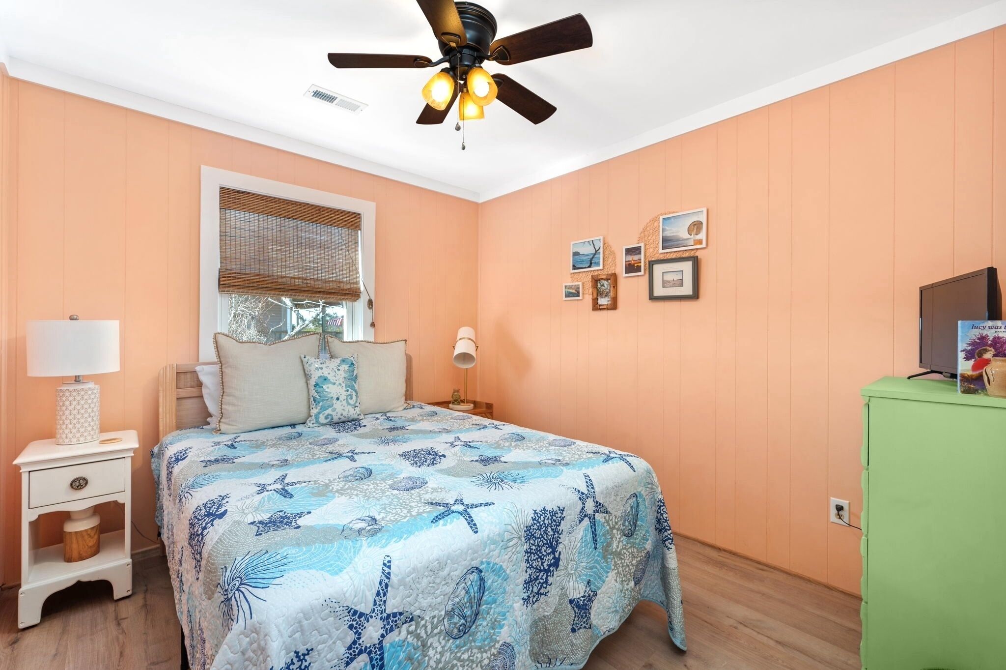 Un dormitorio con cama, mesita de noche, un armario verde, ventilador de techo e imágenes colgadas en la pared.