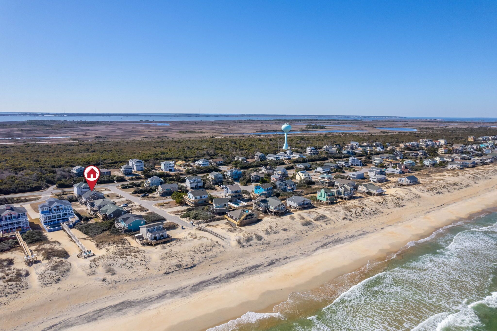 An aerial view of a coastal residential area with houses along the beach, a lighthouse, and a clear sky.