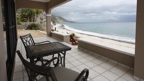 A covered patio with a table and chairs, overlooking a beach with a view of the ocean and distant hills.