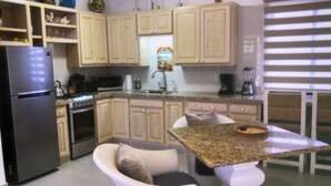 A kitchen with wooden cabinets, a refrigerator, stove, microwave, sink, and a dining area with a chair and table.