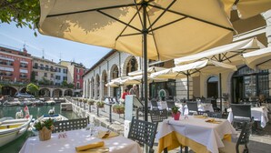 An outdoor dining area with tables and chairs under large umbrellas, overlooking a canal with boats. - Appartamento di Tommaso Central Desenzano w/ A/C sleeps 4, Desenzano Del Garda, Italy (Desenzano Del Garda)