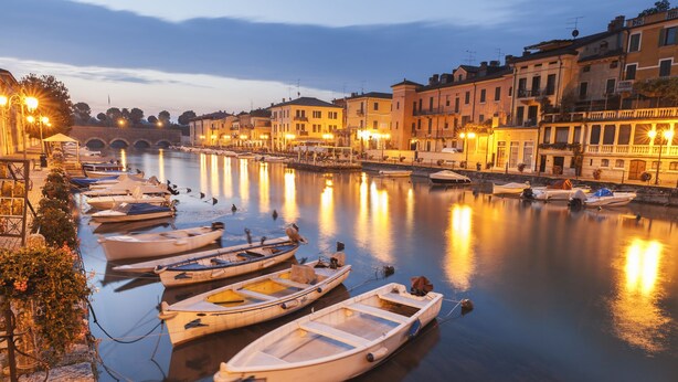 A row of small boats docked along a calm river at dusk, with buildings and a bridge in the background. - Appartamento di Tommaso Central Desenzano w/ A/C sleeps 4, Desenzano Del Garda, Italy (Desenzano Del Garda)