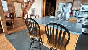 Une cuisine avec des meubles en bois, un réfrigérateur en acier inoxydable, une cuisinière et deux chaises noires disposées autour d’un îlot central en bois.