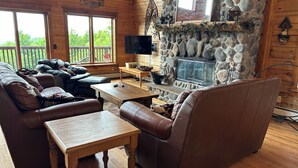 L’intérieur chaleureux d’un chalet avec une cheminée en pierre, des meubles en bois et de grandes fenêtres donnant sur une terrasse.