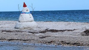 Un muñeco de nieve con sombrero rojo en una playa rocosa con el océano al fondo.