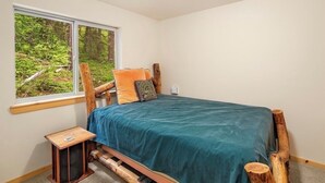 Ein Schlafzimmer mit Bett, das an ein Fenster grenzt, einem kleinen Nachttisch und Blick auf einen Wald.