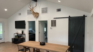 Une pièce spacieuse avec une grande table à manger en bois, des bancs, une tête de cerf accrochée au mur, une porte coulissante de style grange et un panneau d’accueil.