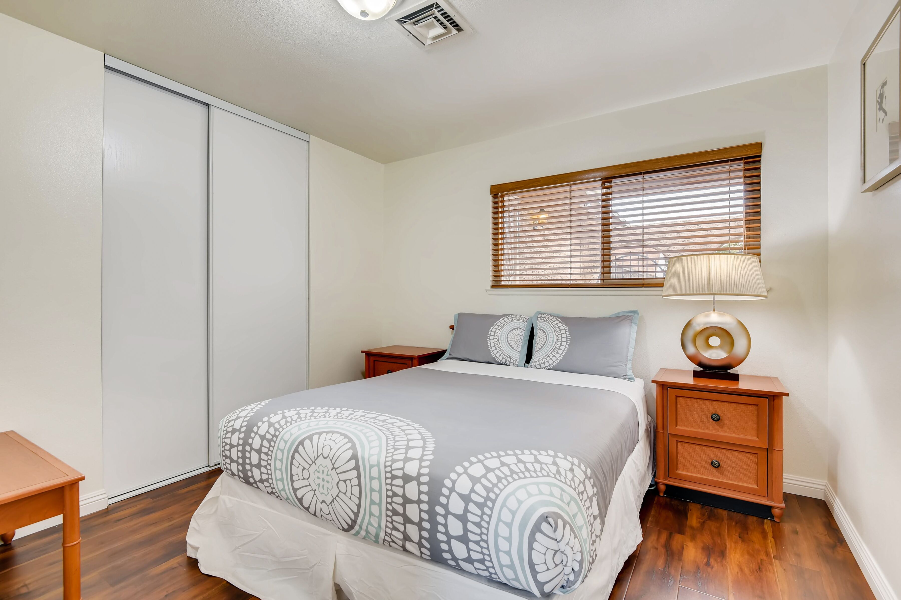 A bedroom with a bed, bedside tables, a nightstand, a window with blinds, and a ceiling vent.